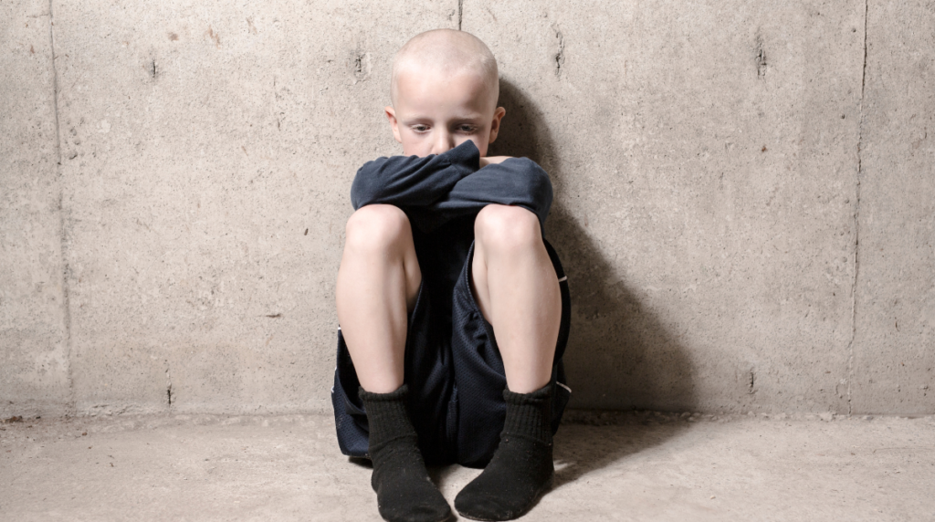 Child showing signs of emotional neglect, sitting alone and withdrawn against concrete wall, demonstrating physical manifestation of emotional isolation